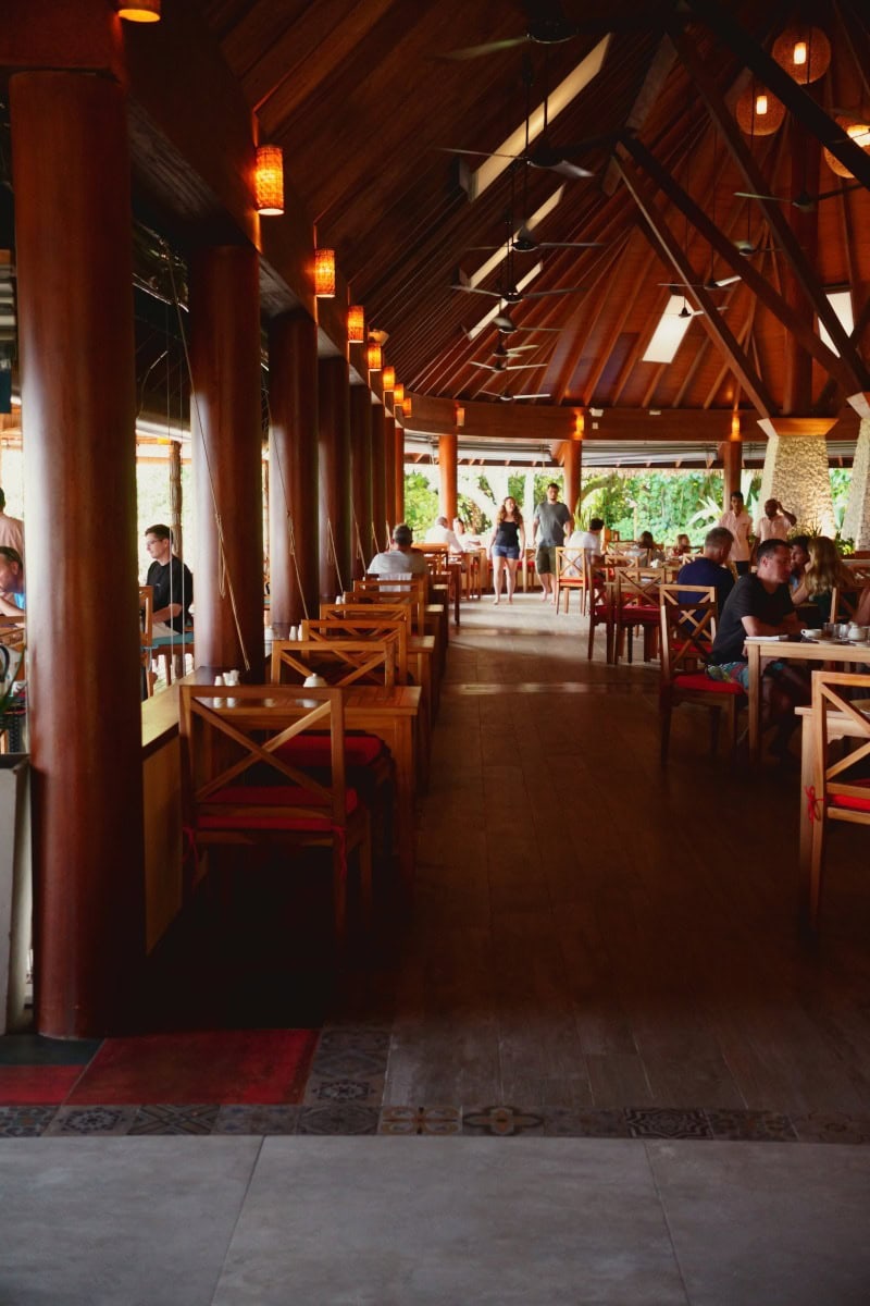 Inside of Vakaru restaurant at Reethi Faru Resort in the Maldives, showing a large wooden pavillion and columns and guests having breakfast