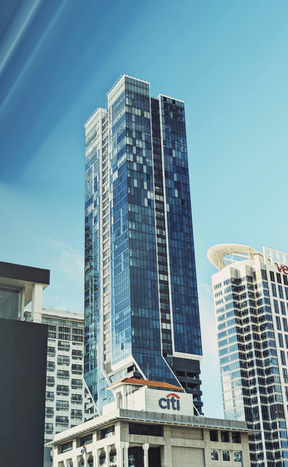 Moderner Glas-Wolkenkratzer mit reflektierender Fassade vor blauem Himmel, flankiert von weiteren Hochhäusern.