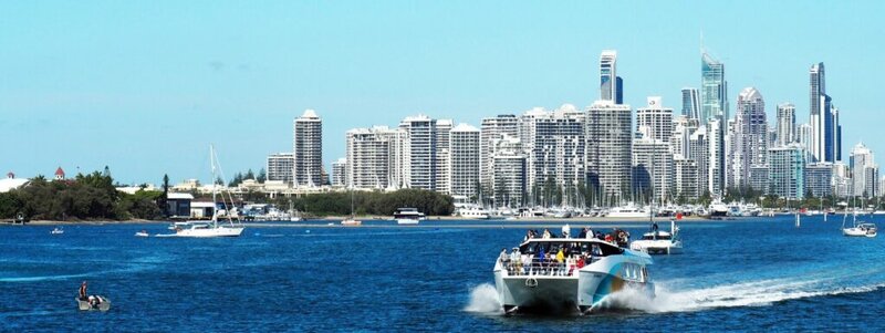 Boot mit Touristen fährt durch blaues Wasser vor der beeindruckenden Skyline einer modernen Küstenstadt.
