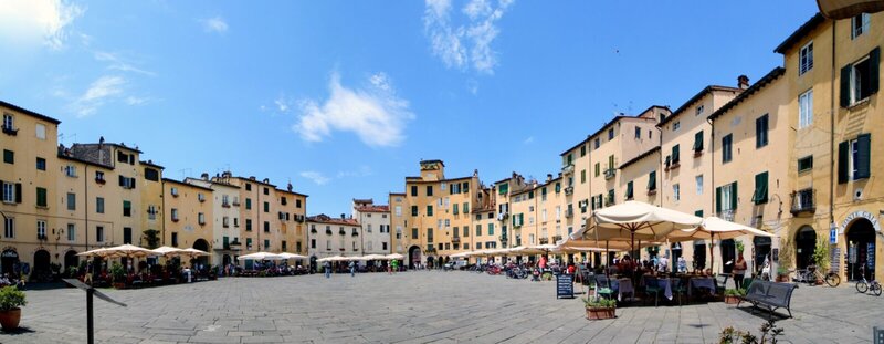 Der Piazza dell’Anfiteatro in Lucca, Italien, ein historischer Platz mit bunten Gebäuden, Cafés und Restaurants unter blauem Himmel.