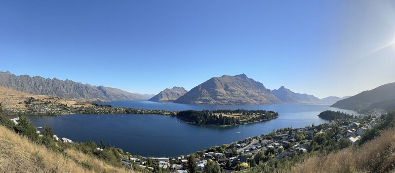 Panoramablick auf den Lake Wakatipu in Queenstown, Neuseeland, mit der Halbinsel Kelvin Heights, den Remarkables-Bergen und klar blauem Himmel.