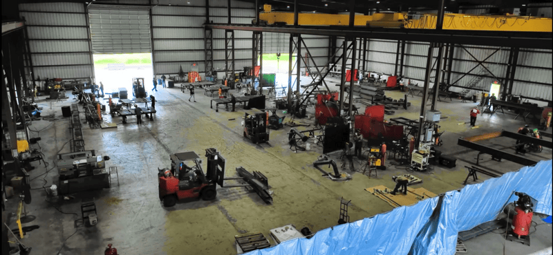 Interior of WeldIT's 35,000 sq ft metal fabrication shop in Houston with overhead bridge cranes, welding stations, forklifts, and structural steel in production