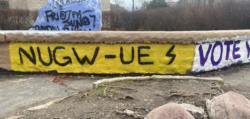 Bench in front of The Rock painted with the letters N U G W dash U E S and the word "vote."