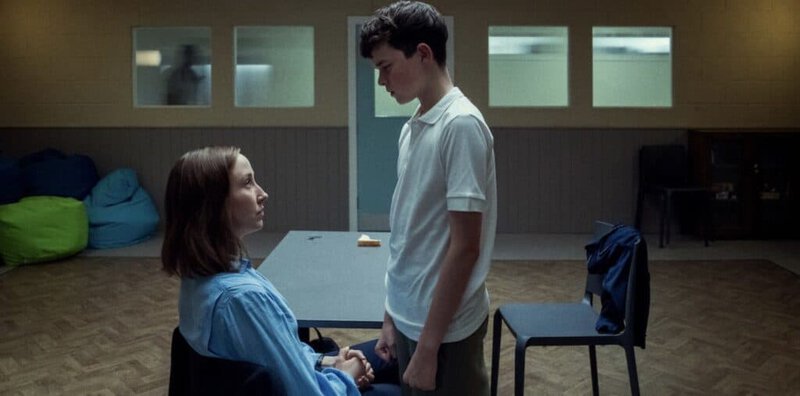 Still from Adolescence showing a teenage boy standing in front of a woman in a dimly lit institutional room, capturing a moment of confrontation and empathy.