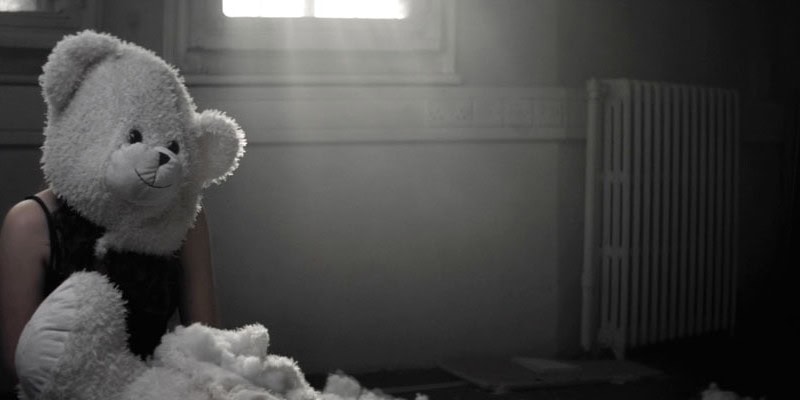 Cinematic music video production shot featuring a surreal, dark narrative scene with a woman in a teddy bear mask in a derelict room.