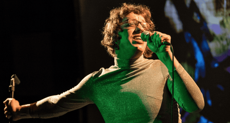 Harry Josephine Giles with short curly hair and glasses passionately sings into a microphone on stage, wearing a shimmery long-sleeve top, with dramatic lighting and a colorful background.