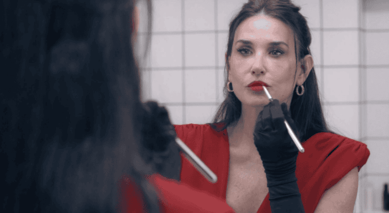 Still from The Substance of a woman in red dress applying lipstick in front of a mirror, reflecting themes of identity and duality.
