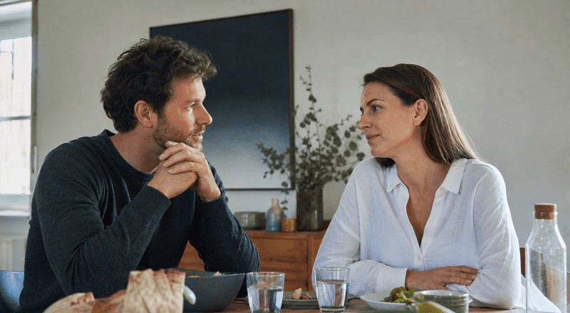Man and woman sit across from each other at a dining table, engaging in a thoughtful conversation over a meal with bread and bowls nearby.
