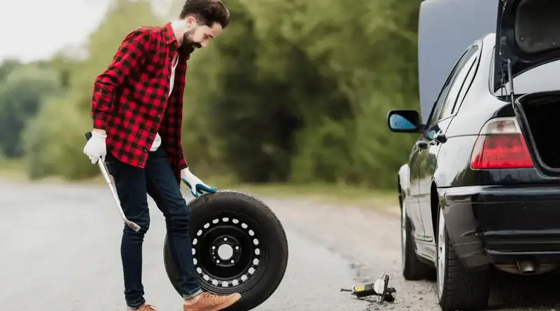 Homem jovem trocando o pneu do carro na estrada ilustrando o artigo sobre pneu furado