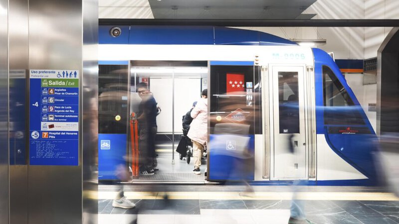 Un tren del Metro de Madrid en una estación, con personas entrando y saliendo, y señalización visible en la pared.