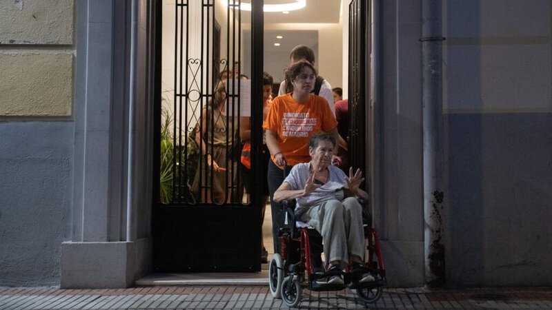 Una mujer mayor en silla de ruedas es acompañada por una persona mientras sale de un edificio, con un grupo de personas al fo