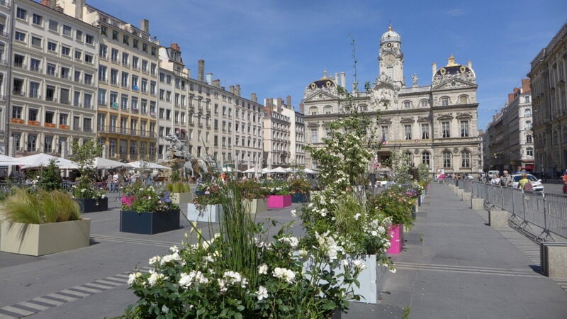 Historischer Stadtplatz mit Blumenarrangements, Cafés und beeindruckender Architektur bei sonnigem Wetter.
