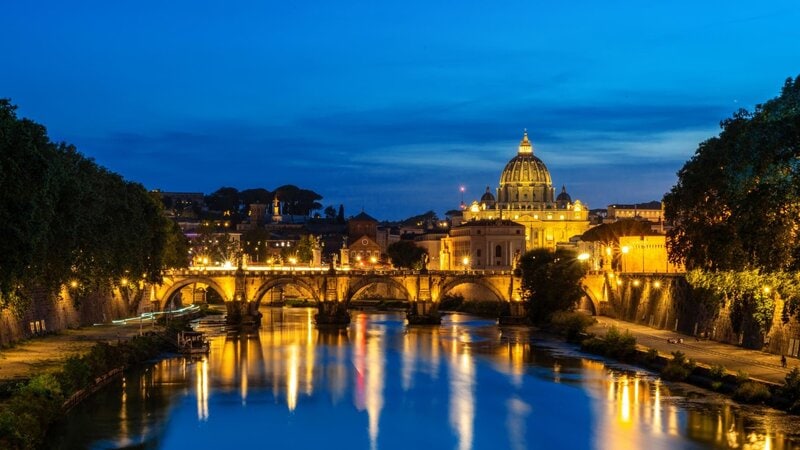Beleuchteter Petersdom und Engelsbrücke in Rom bei Nacht, mit Spiegelung im Tiber.