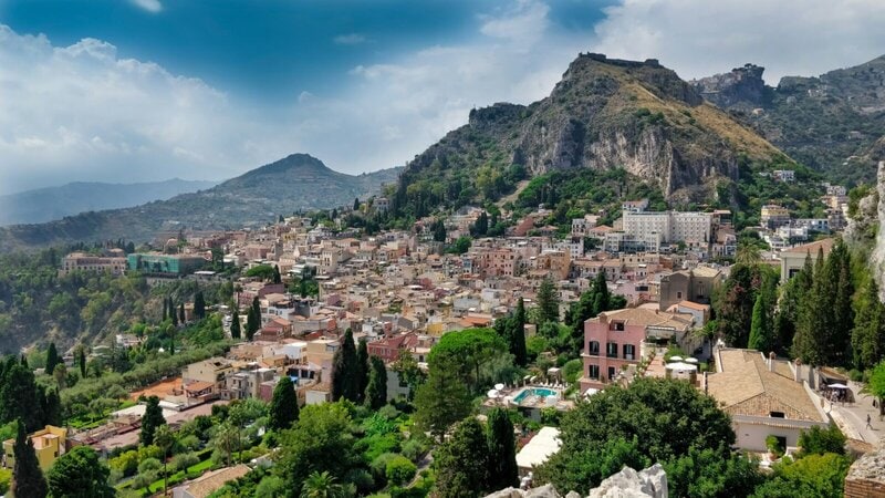 Panoramablick auf die malerische Stadt Taormina auf Sizilien, umgeben von grünen Hügeln und historischen Gebäuden.