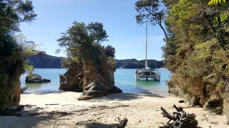 Idyllische Strandbucht mit Felsen und tropischer Vegetation im Abel Tasman Nationalpark, Neuseeland. Ein Katamaran ankert im türkisblauen Wasser.