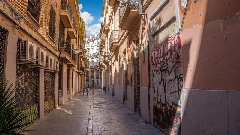 Schmale Gasse in Valencia mit historischen Gebäuden, Graffiti und einem Fußgänger mit Fahrrad.