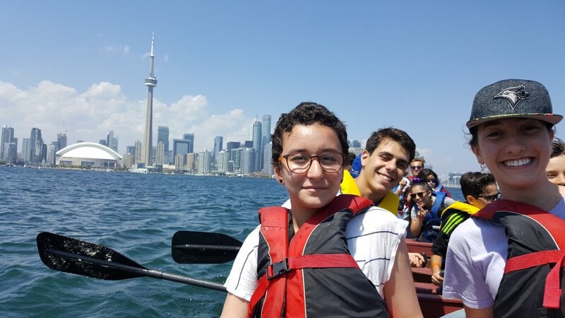 Eine Gruppe junger Menschen beim Kanufahren mit Blick auf die Skyline von Toronto, inklusive CN Tower und Rogers Centre.