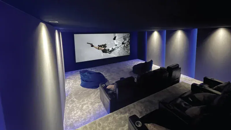 Experience a modern home theatre room with dark walls, recliner seats, a large screen showcasing an action film, soft carpet flooring, and ambient blue lighting—an ideal home cinema sample in our Surrey showroom.