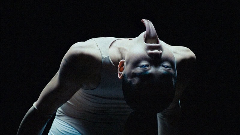 Contortionist dancer in Bonnie Banane’s ‘Hoes of Na’ bending backward in a dark, surreal setting with an elongated tongue effect.
