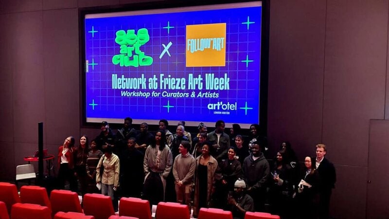 A group of people poses in front of a large screen in a theater. The screen reads, Network at Frieze Art Week: Workshop for Curators & Artists, with event logos, artotel London Battersea Power Station, and FOLLOW.ART featured.