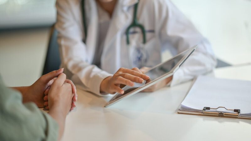 A doctor in a white coat uses a tablet while talking with a patient; a clipboard and papers are on the table.