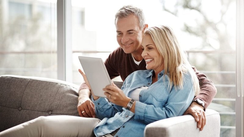 Darstellung von zwei Personen, die auf dem Sofa sitzen und lächelnd in ein Tablet gucken.