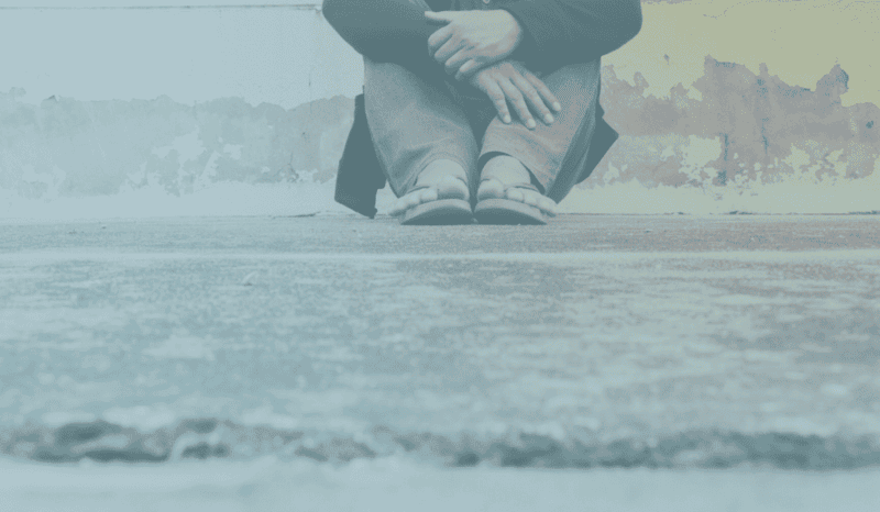 Beitragsbild zu Depression erkennen: Eine Person, deren Kopf nicht zu sehen ist, sitzt mit angewinkelten Beinen auf dem Boden und hat die Arme um die Knie geschlungen. Es scheint, dass die Person depressiv, traurig oder nachdenklich ist. Der Hintergrund ist eine Wand mit abblätternder Farbe, was ein Gefühl von Vernachlässigung vermittelt. Die Farbpalette des Bildes ist kalt und gedämpft, was eine Stimmung der Einsamkeit und Traurigkeit unterstreicht. Der allgemeine Eindruck ist einer der Melancholie oder des emotionalen Rückzugs.