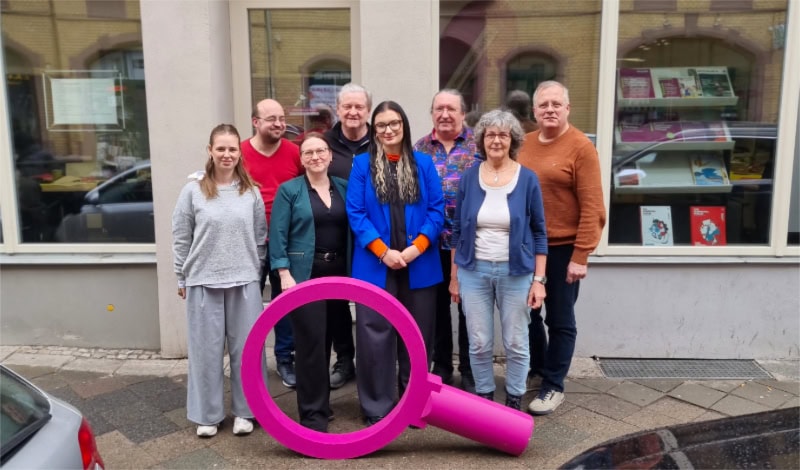 Eine Gruppe von Menschen steht eng beieinander auf einem Gehweg vor einem Gebäude mit großen Fenstern. Im Vordergrund befindet sich eine überdimensionale, beerenfarbige Lupe, die aufrecht auf dem Boden steht. Die Personen sind unterschiedlich gekleidet, darunter Jacken, Pullover und eine auffällige blaue Jacke. Im Hintergrund spiegeln sich geparkte Autos in den Fenstern. Die Szene zeigt den aktuellen Vorstand des Kommunalpolitischen Bildungsvereins Die Thüringengestalter.