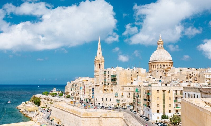 Blick auf die Skyline von Valletta, Malta, mit historischen Gebäuden, der Kuppel der Karmeliterkirche und dem Mittelmeer im Hintergrund.