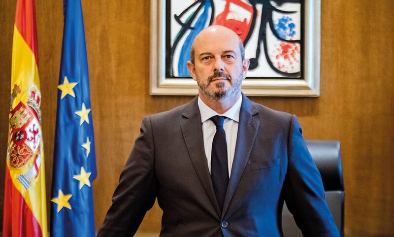 Pedro Rollán en un entorno oficial, con banderas española y europea, y una pintura abstracta en la pared detrás.