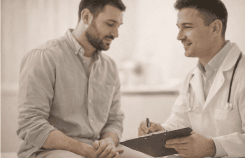 A doctor in a white coat holding a clipboard speaks with a seated male patient during a medical consultation.