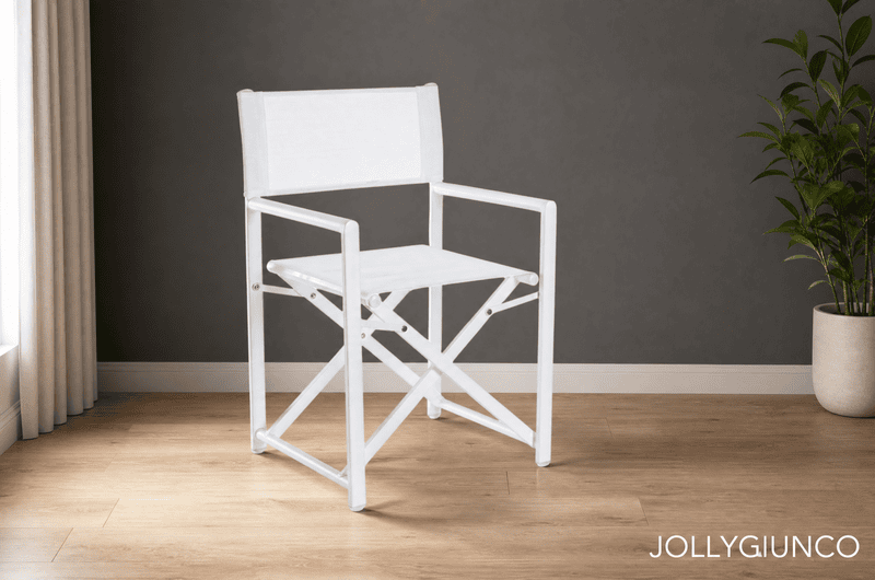 White director-style folding chair on a light wood floor against a dark gray wall, with beige curtains on the left and a tall plant in a pot on the right; watermark reads JOLLYGIUNCO at bottom right.