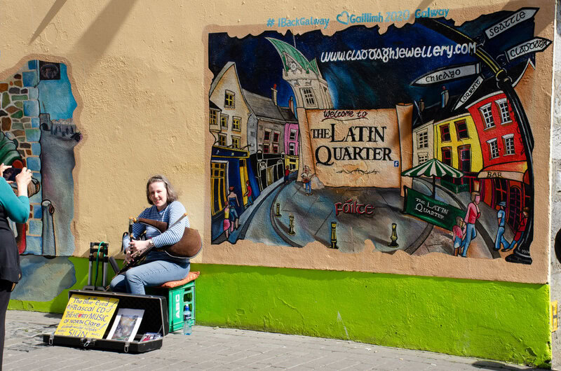 Straßenmusikerin spielt Uilleann Pipes vor einer bunten Wandmalerei im Latin Quarter, Galway.