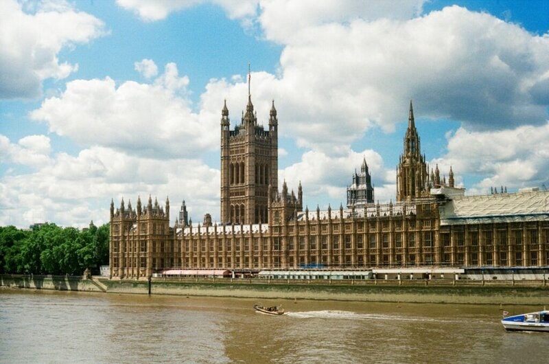 Das Houses of Parliament in London mit der Themse im Vordergrund und blauem Himmel mit weißen Wolken im Hintergrund.
