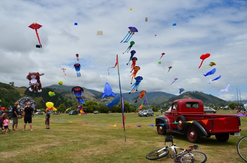 Bunte Drachen steigen in den Himmel bei einem Drachenfestival auf einer grünen Wiese mit Bergen im Hintergrund. Ein roter Oldtimer-Truck und Zuschauer sind ebenfalls zu sehen.