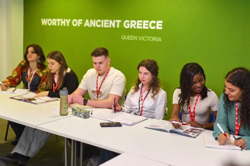 Gruppe von internationalen Studierenden im Klassenzimmer mit grünem Hintergrund und dem Zitat von Queen Victoria.