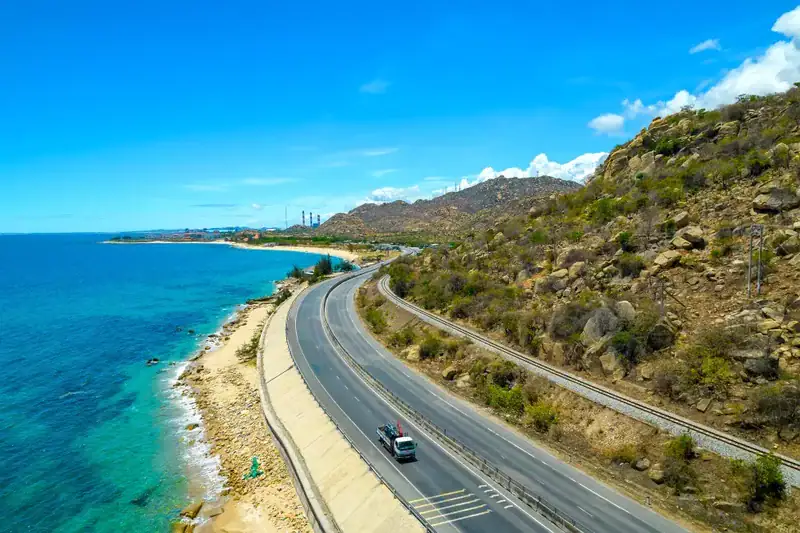 Rodovia costeira com caminhão branco, mar azul e colinas rochosas sob céu azul claro. Conteúdo sobre pagar pedágio online no Litoral Norte