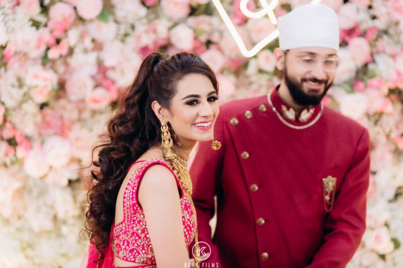 Phillip and Naina smile in front of a pink and white flower wall at their wedding at Namdhari Sangat, Thailand