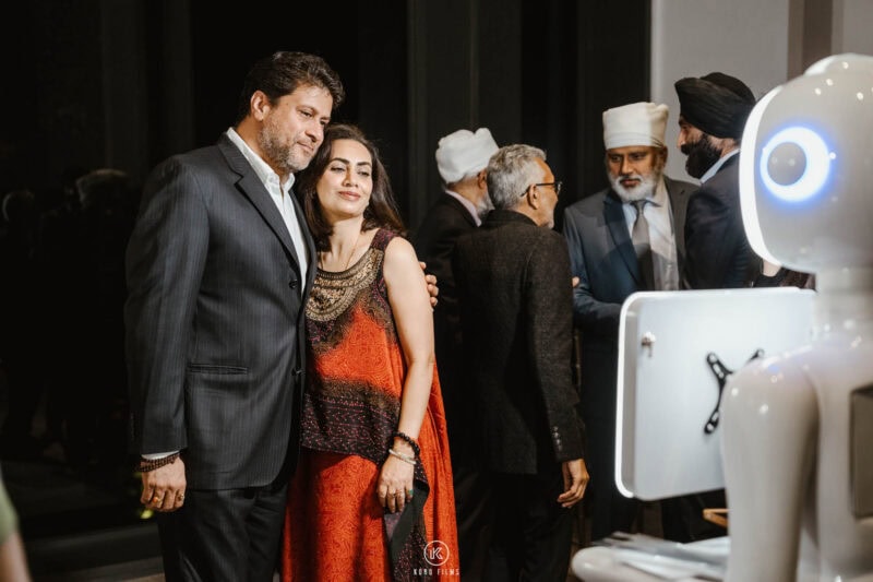 Couple in festive outfits posing in front of the robot photo booth at a wedding