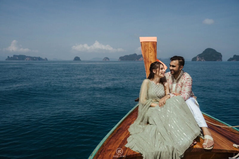 Bride in a sage-green lehenga and groom in a printed cream outfit sit on the bow of a long-tail boat with limestone islands behind, pre-wedding shoot at Ko Hong, Phang Nga