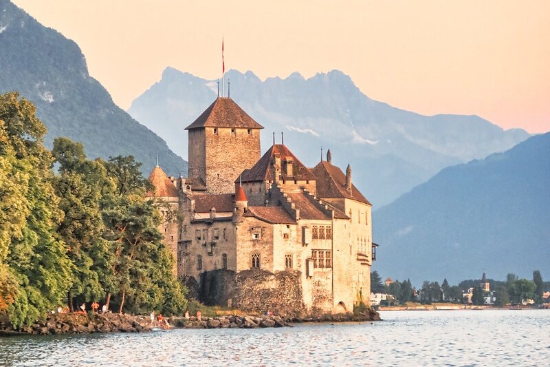 Historisches Schloss am Seeufer mit malerischer Bergkulisse bei Sonnenuntergang.