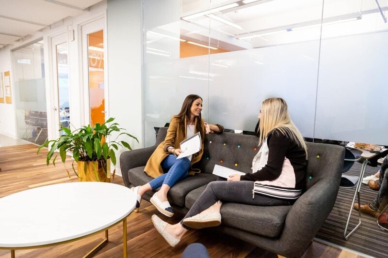 Zwei junge Frauen unterhalten sich in einer modernen Lounge-Bereich einer Bildungseinrichtung mit entspanntem Ambiente.