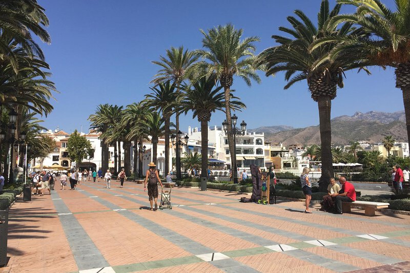 Menschen spazieren auf dem palmengesäumten Platz Balcón de Europa in Nerja mit Blick auf die umliegenden Berge