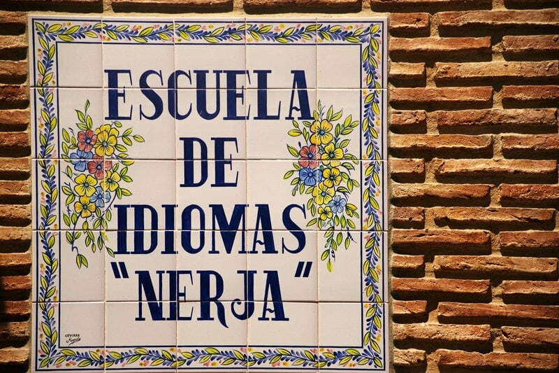 Keramikschild mit floraler Verzierung und Aufschrift „Escuela de Idiomas Nerja“ an einer Ziegelwand