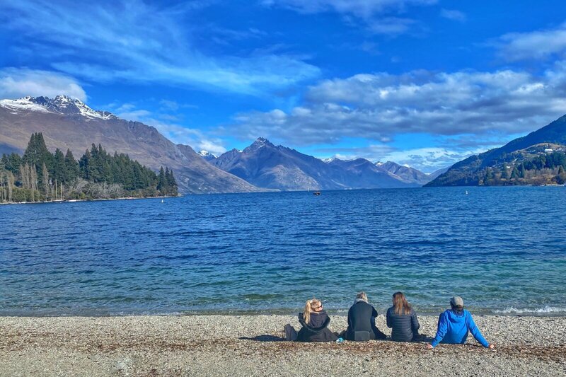 Vier Personen sitzen am Ufer des Lake Wakatipu in Queenstown, Neuseeland, mit Blick auf die schneebedeckten Berge unter einem blauen Himmel.