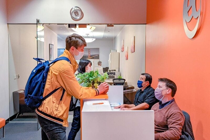 Ein junger Mann mit Rucksack spricht mit Mitarbeitern an einer Rezeption in einem modernen Büro, alle tragen Schutzmasken.