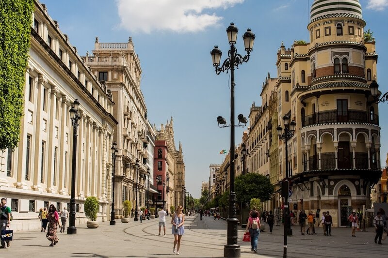 Historische Straße in Sevilla mit Fußgängern, klassischen Straßenlaternen und markanten Gebäuden im andalusischen Stil.