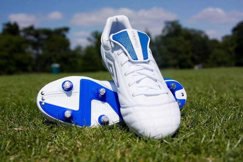 Weiße Fußballschuhe mit blauen Akzenten und Stollen auf einem grünen Rasen unter blauem Himmel.