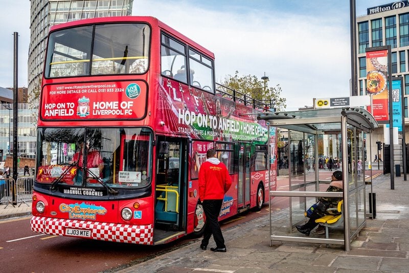 Roter Liverpool City Explorer Doppeldeckerbus mit Werbung für Anfield-Touren an einer Bushaltestelle.