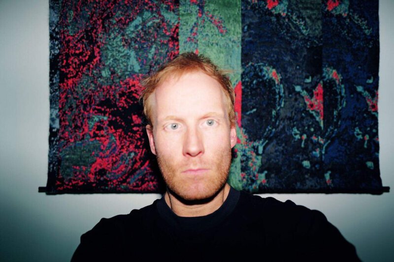 A man with light skin and reddish hair stares directly at the camera, illuminated by a flash. Behind him is a colorful, abstract tapestry with green, red, and dark blue patterns.